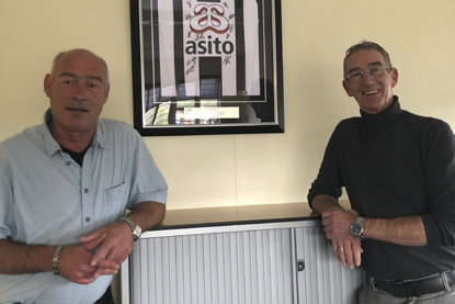 Afdelingshoofd Erwin Teiwes en vestigingsdirecteur Ton Golstein poseren. Tussen hen in hangt een ingelijst shirt van voetbalclub Heracles Almelo. De sponsor op het shirt is schoonmaakbedrijf Asito, een netwerkpartner van PI Almelo.